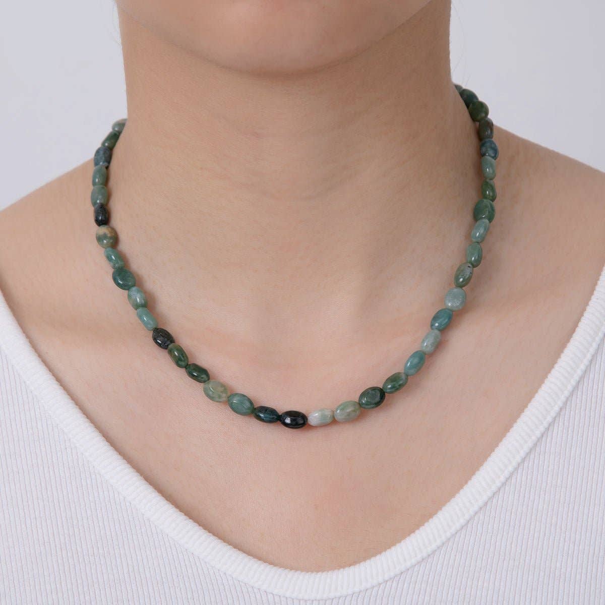 A beaded necklace made of polished oval beads in shades of green, teal, and dark charcoal, worn on a model's neck over a white ribbed top.