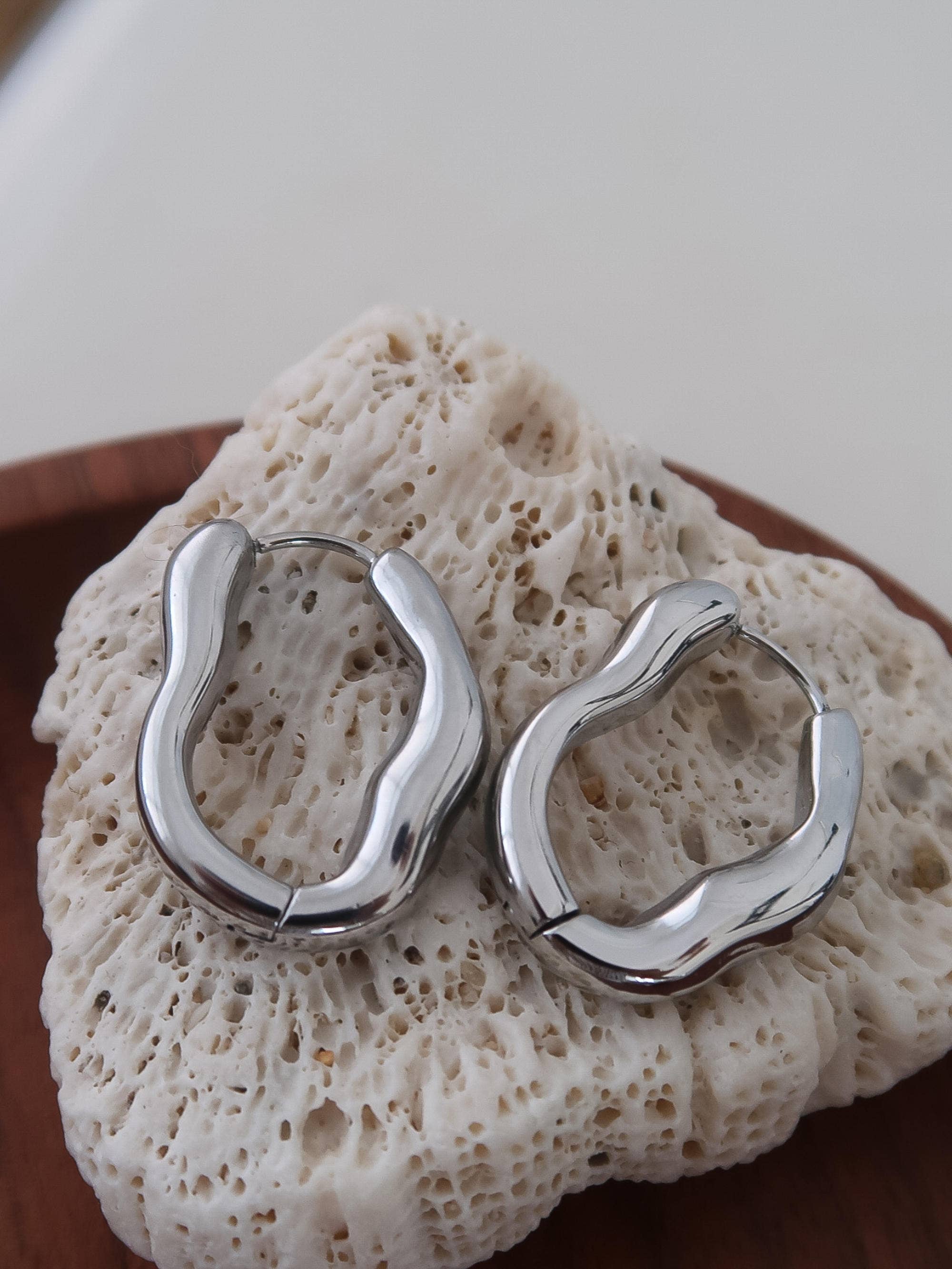 Wavy oval silver hoop earrings on a textured stone surface
