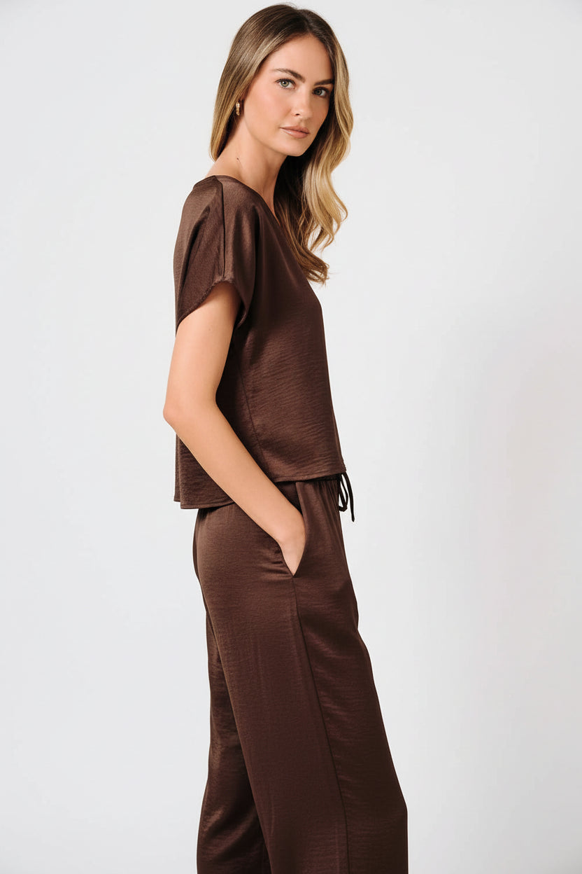 A woman modeling a brown crinkle satin top with dolman short sleeve and V-neckline against a solid beige background.
