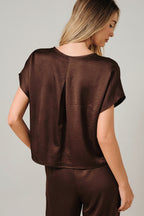 Back view of a woman modeling a brown crinkle satin top with dolman short sleeve against a solid beige background.