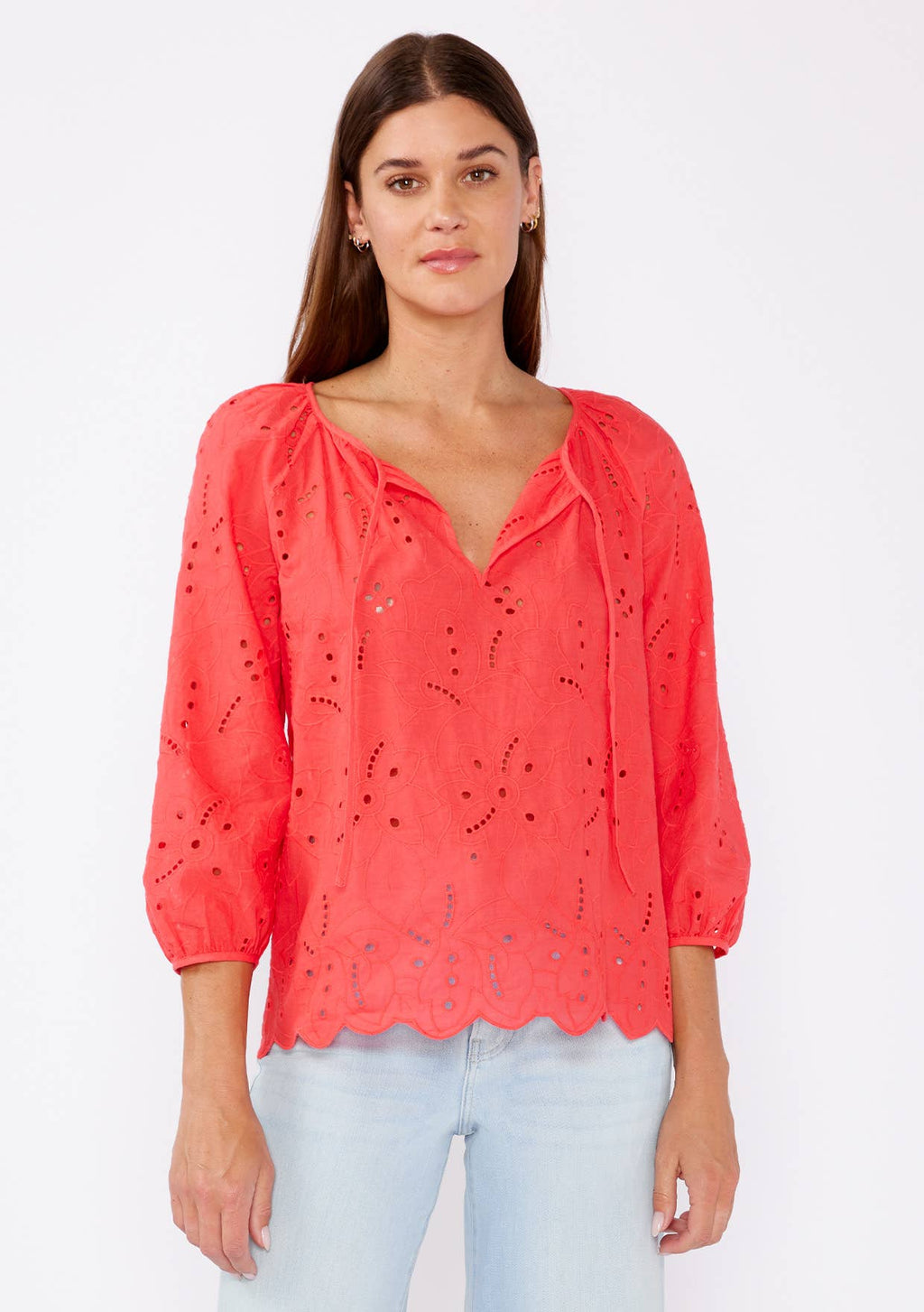 Woman wearing a coral eyelet top with scalloped hemline and raglan sleeves and light blue pants on a white background
