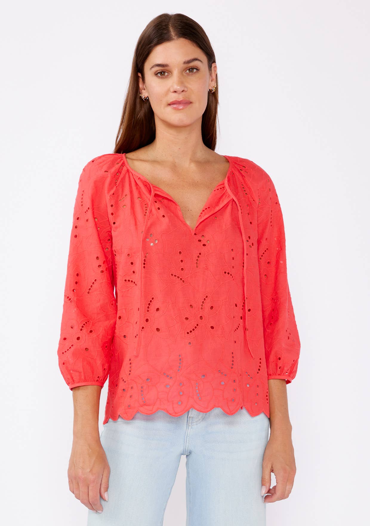 Woman wearing a coral eyelet top with scalloped hemline and raglan sleeves and light blue pants on a white background