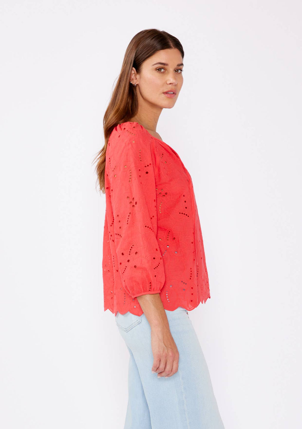 Woman wearing a coral eyelet top with scalloped hemline and raglan sleeves and light blue pants on a white background