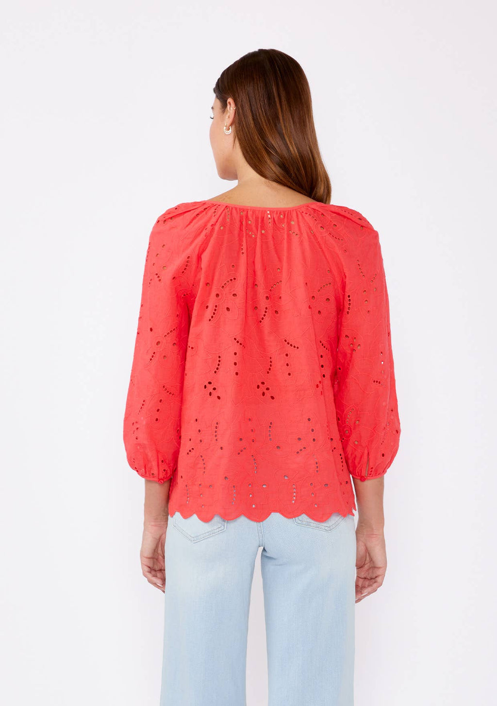 Woman wearing a coral eyelet top with scalloped hemline and raglan sleeves and light blue pants on a white background