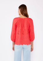 Woman wearing a coral eyelet top with scalloped hemline and raglan sleeves and light blue pants on a white background