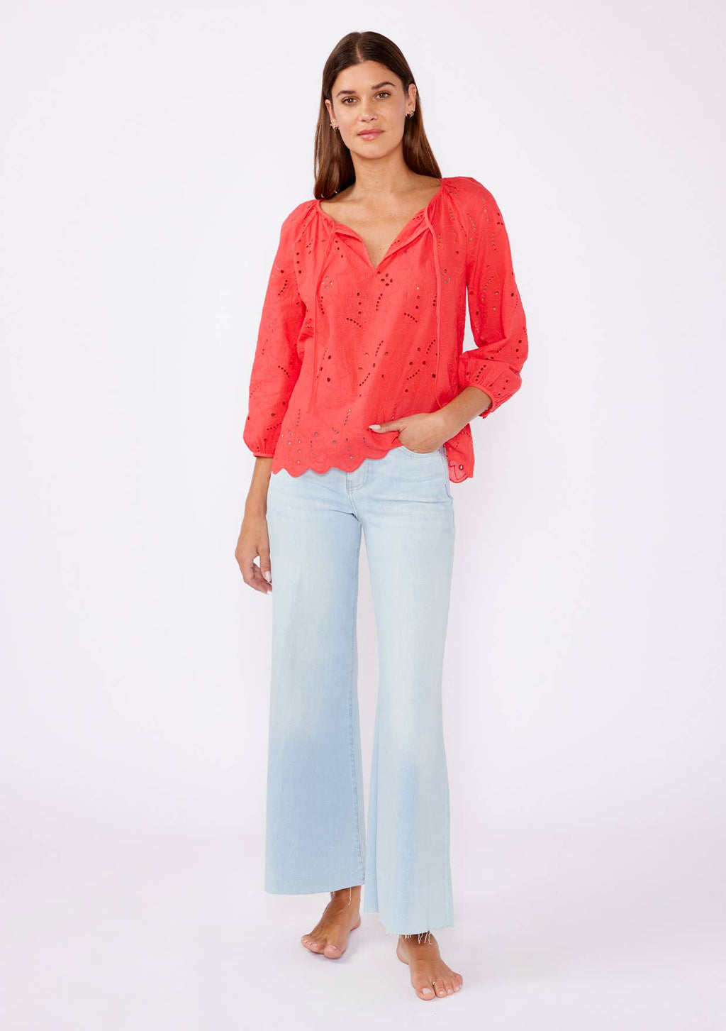 Woman wearing a coral eyelet top with scalloped hemline and raglan sleeves and light blue pants on a white background