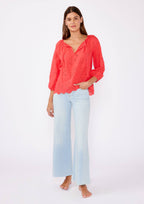 Woman wearing a coral eyelet top with scalloped hemline and raglan sleeves and light blue pants on a white background