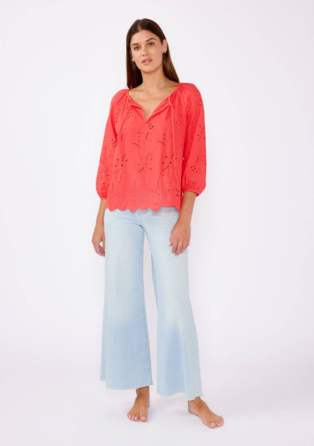 Woman wearing a coral eyelet top with scalloped hemline and raglan sleeves and light blue pants on a white background