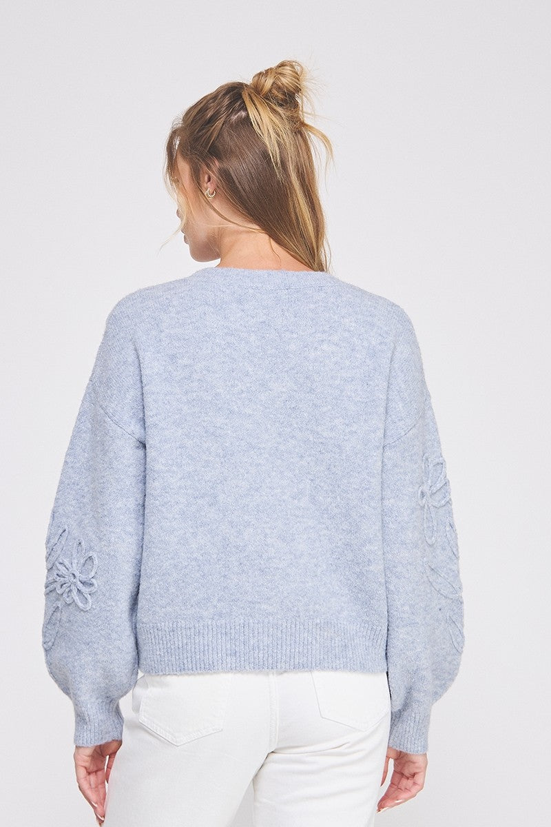 Woman showing the back of a  heather blue knit sweater with floral embroidery on a white background
