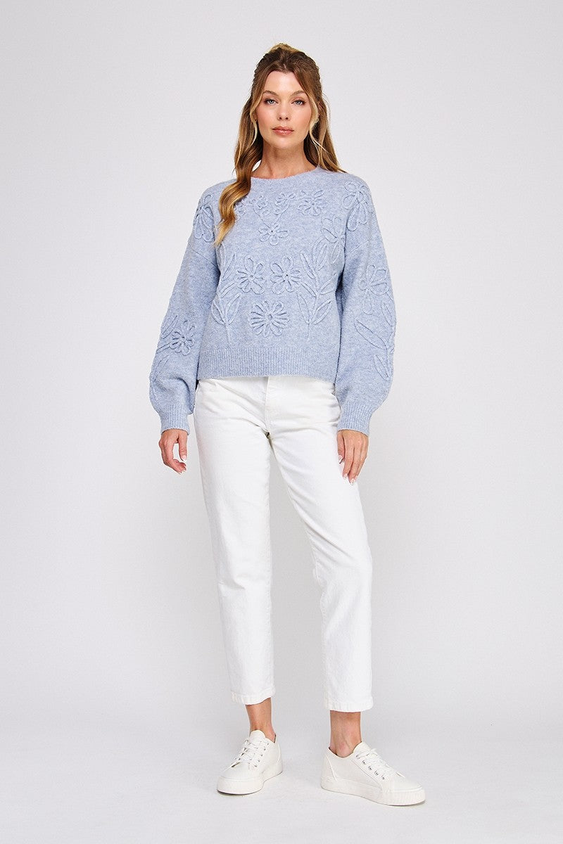 Woman wearing a heather blue knit sweater with floral embroidery and white jeans on a white background