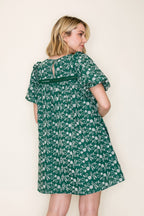 Back view of a woman modeling a short puff sleeve green dress with a white embroidered design, standing against a plain off-white background.