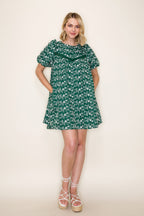 A woman modeling a short puff sleeve green dress with a white embroidered design, standing against a plain off-white background.