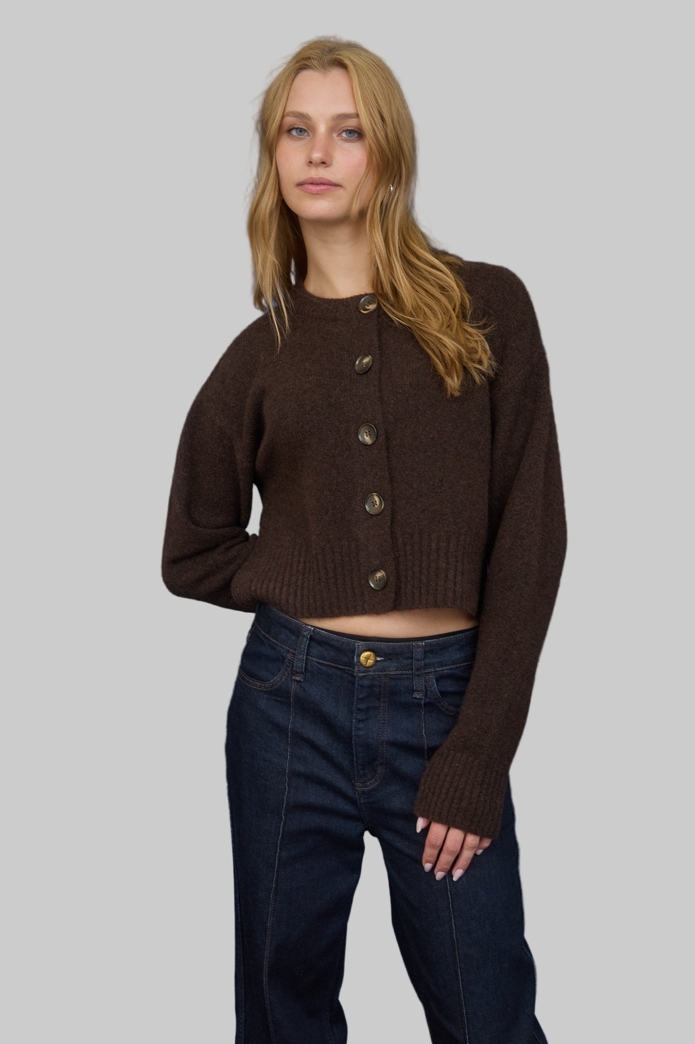 Woman wearing a brown knitted  button front cardigan and dark jeans against a gray background