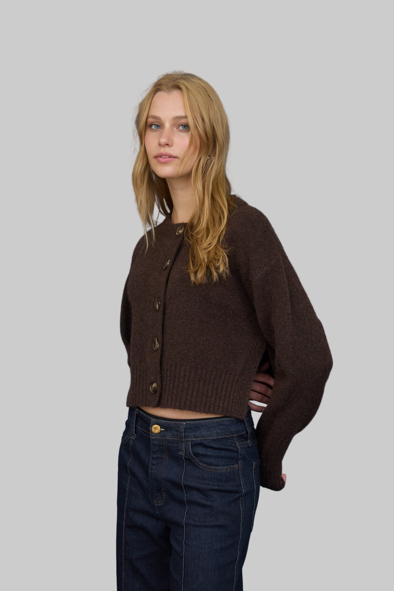 Woman wearing a brown button front cardigan and dark jeans against a gray background