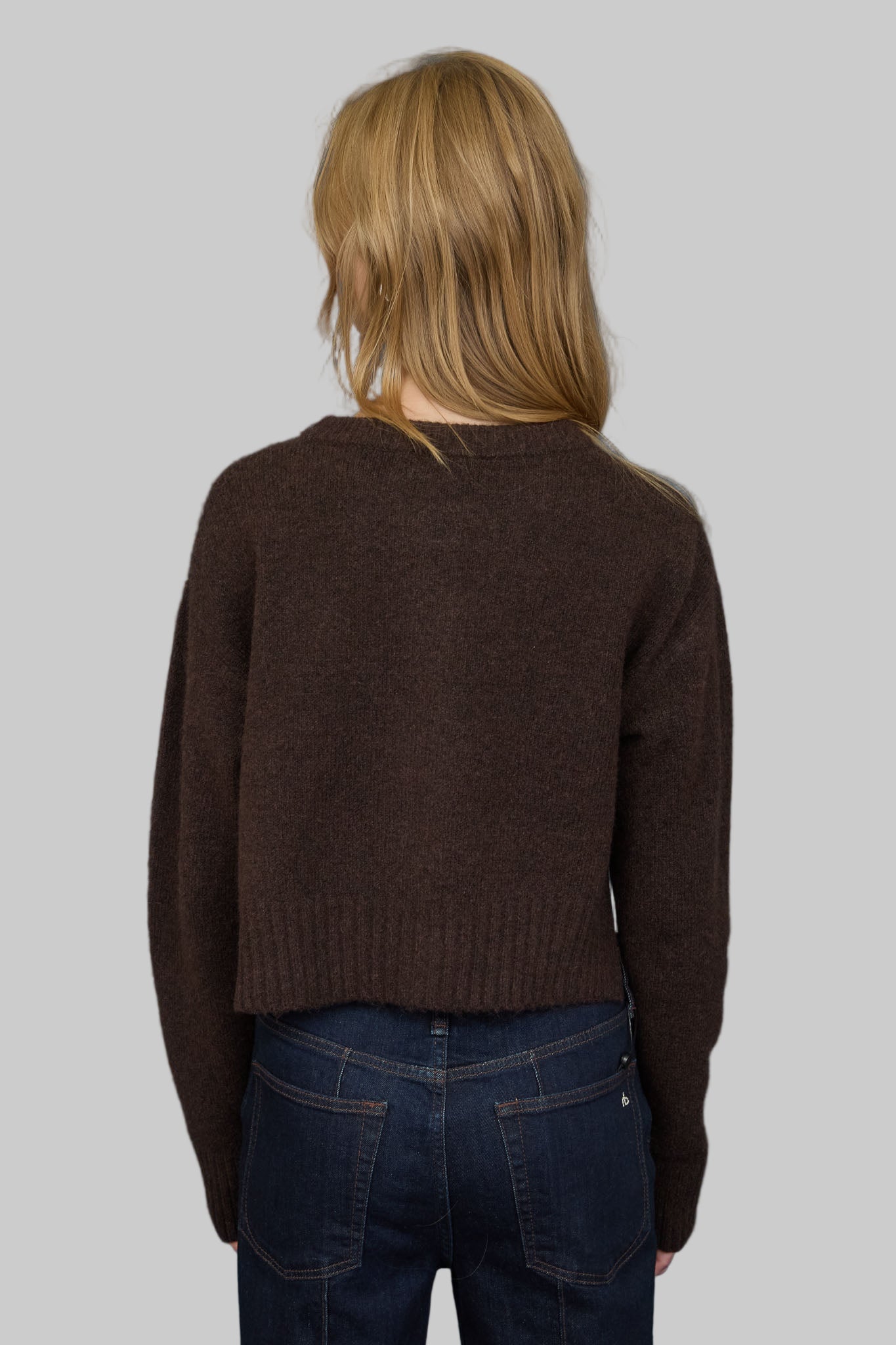 Woman showing back view of a brown sweater and blue jeans against a gray background