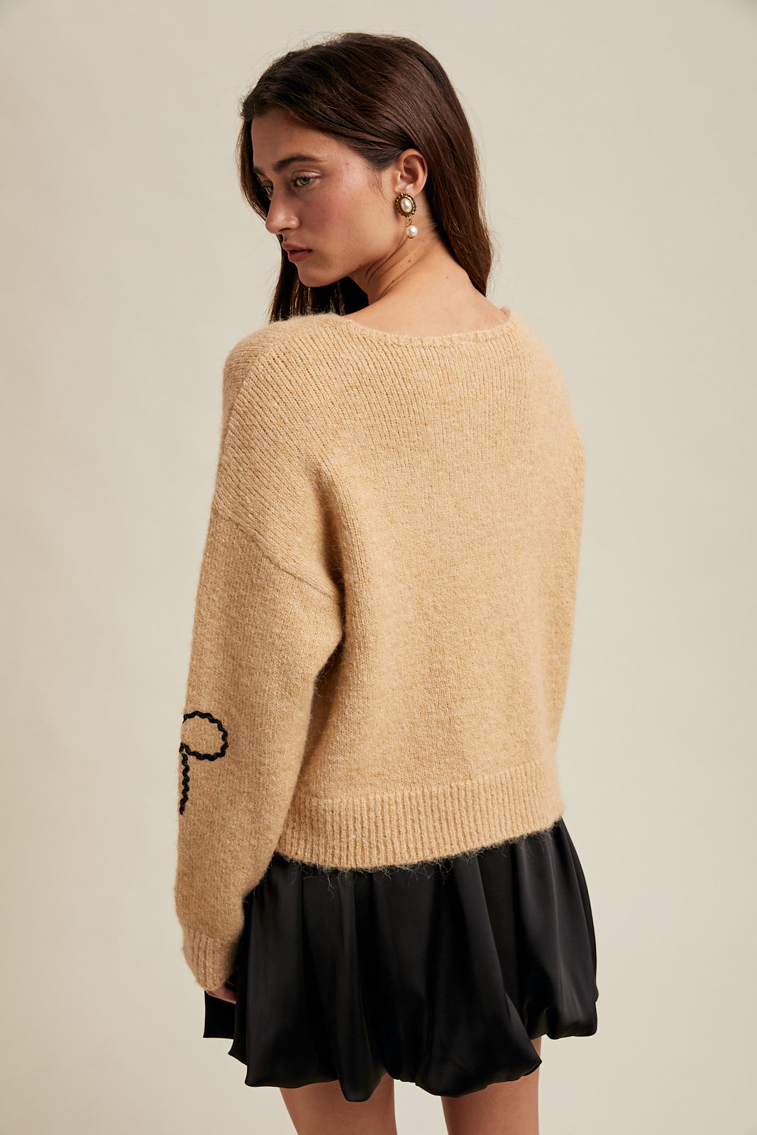 Back view of a woman wearing a taupe sweater with black rick rack bow designs and black skirt on a neutral background