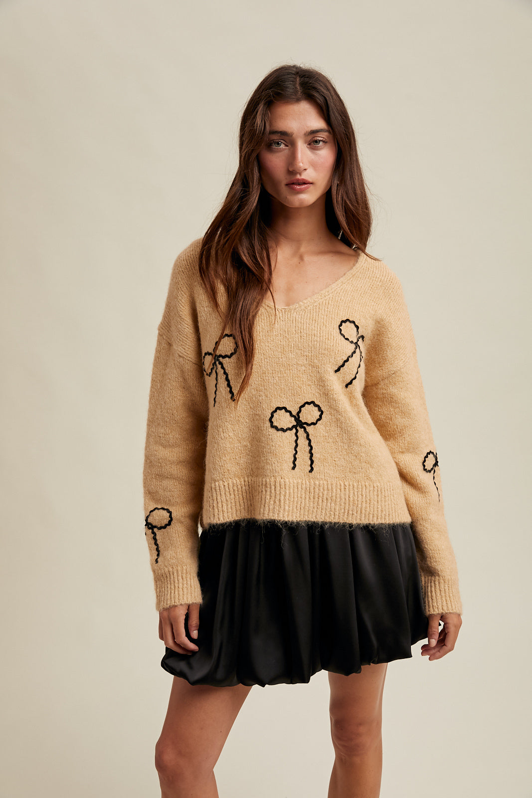Woman wearing a taupe sweater with black rick rack bow designs and black skirt on a neutral background