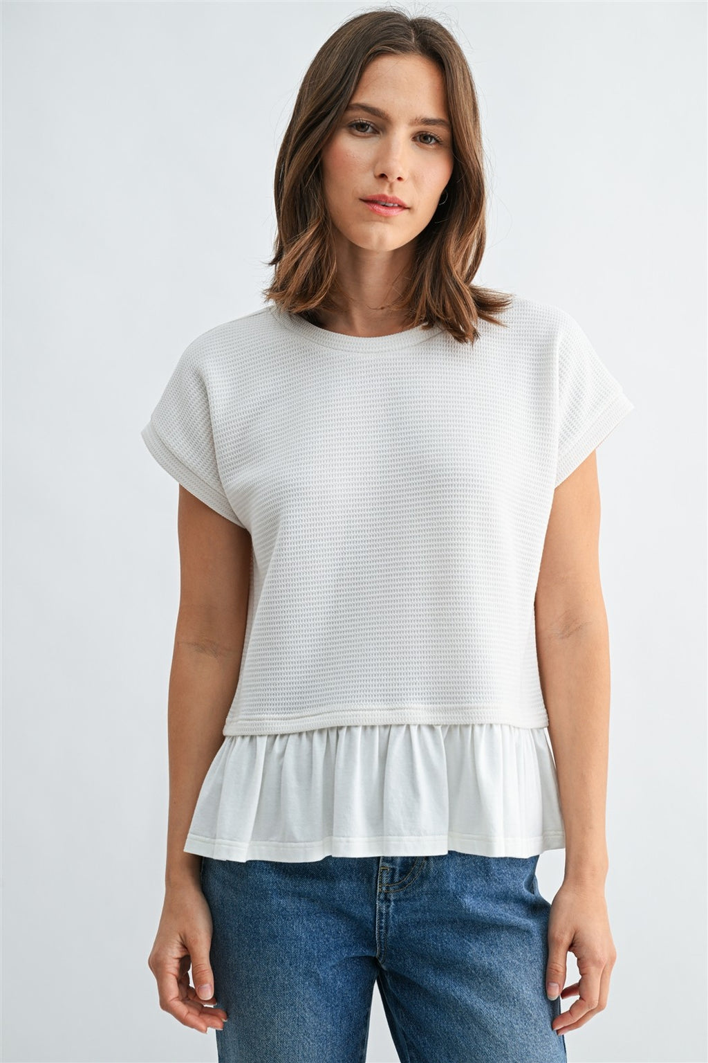 Woman wearing a white knit top with a ruffled bottom and blue jeans on a light gray background