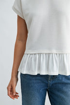 Close up of a woman wearing a white knit textured top with a ruffled bottom and blue jeans on a light gray background