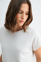 Woman wearing a white textured knit top with a ruffled bottom and blue jeans on a light gray background