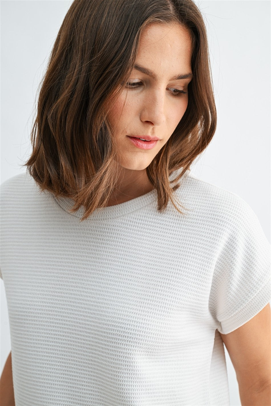 Woman wearing a white textured knit top with a ruffled bottom and blue jeans on a light gray background