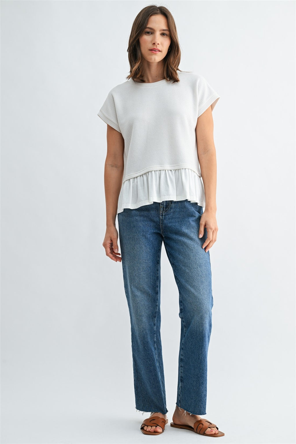 Woman wearing a white knit textured top with a ruffled bottom and blue jeans on a light gray background