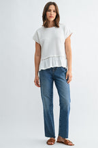 Woman wearing a white knit textured top with a ruffled bottom and blue jeans on a light gray background