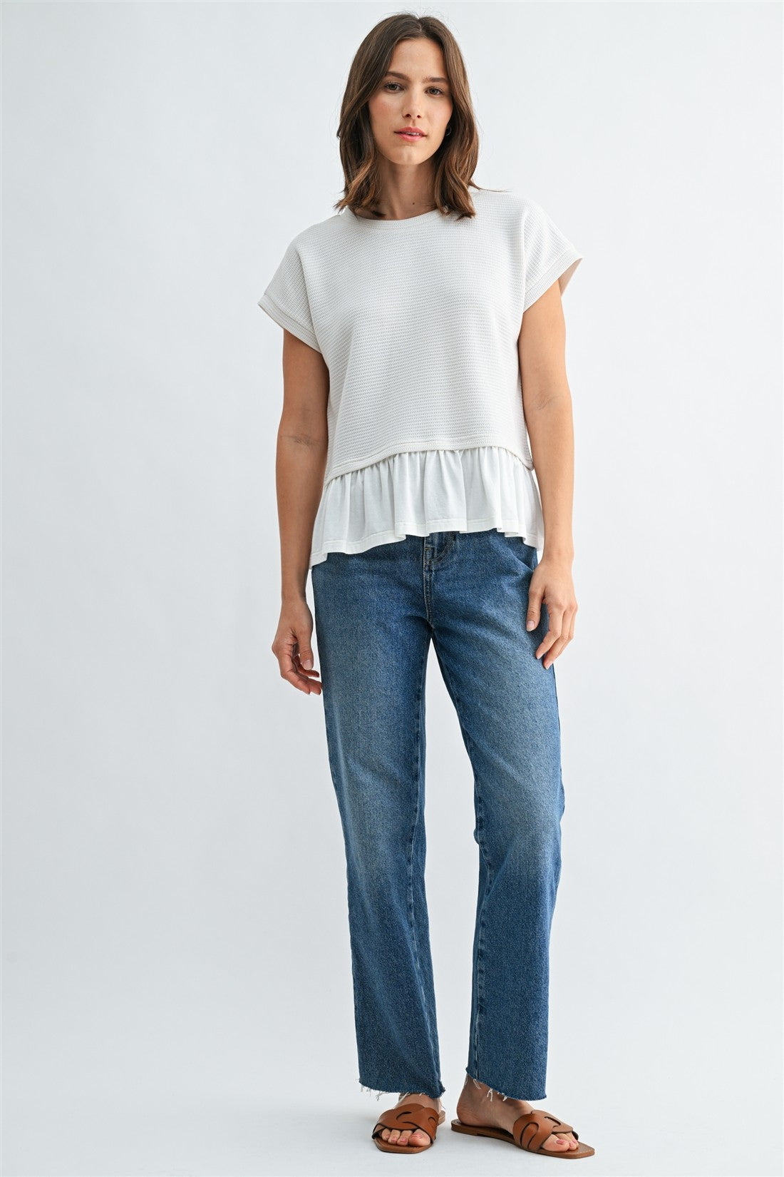 Woman wearing a white knit textured top with a ruffled bottom and blue jeans on a light gray background