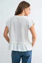 Back view of a woman wearing a white knit textured top with a ruffled bottom and blue jeans on a light gray background