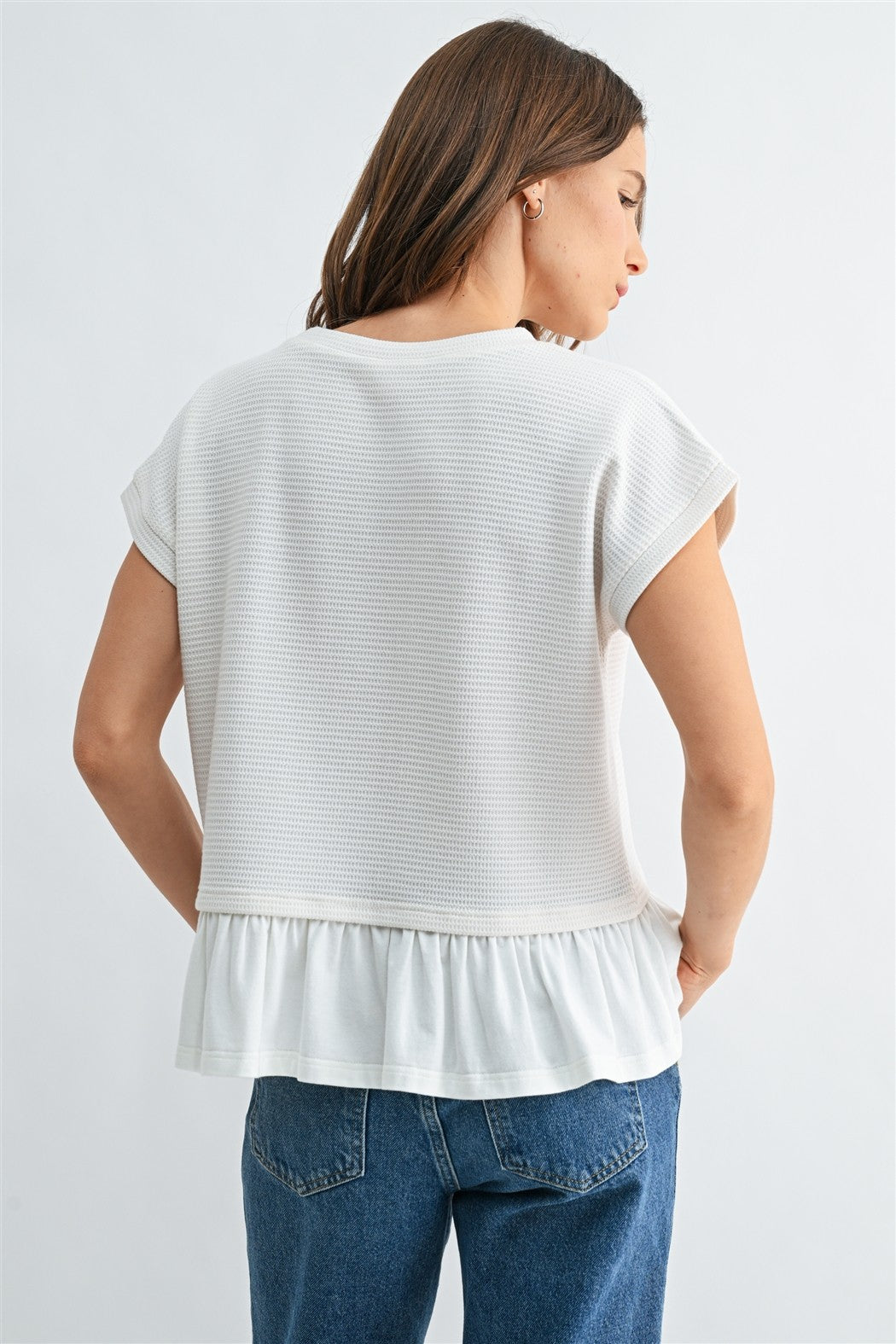 Back view of a woman wearing a white knit textured top with a ruffled bottom and blue jeans on a light gray background