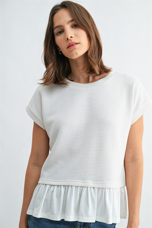 Woman wearing a white knit tpextured top with a ruffled bottom and blue jeans on a light gray background