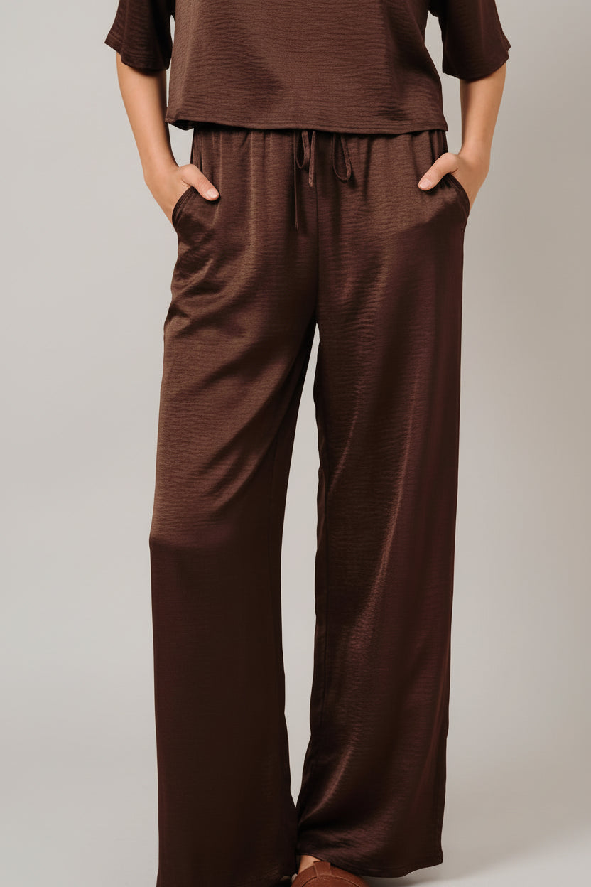 A woman modeling brown crinkle fabric wide leg pants with side pockets and elastic drawstring waistband against a solid beige background.