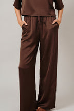 A woman modeling brown crinkle fabric wide leg pants with side pockets and elastic drawstring waistband against a solid beige background.