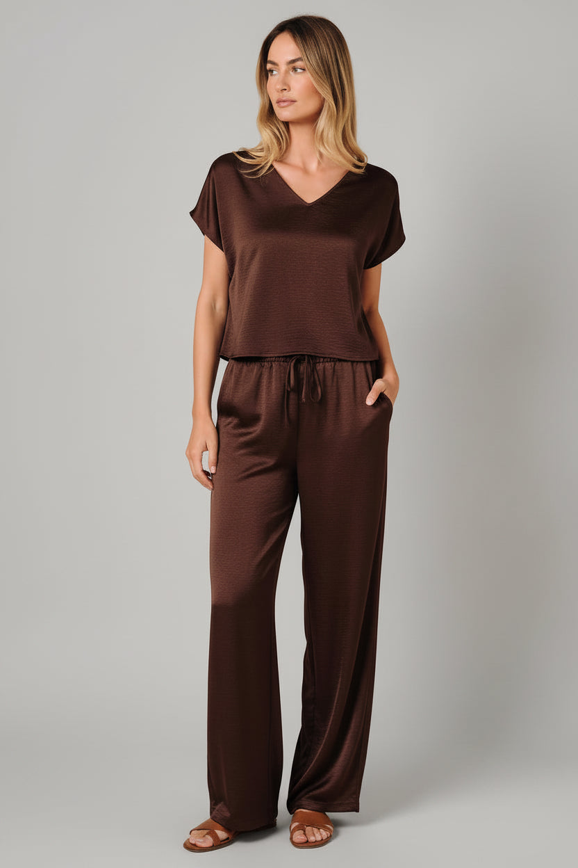 A woman modeling a brown crinkle fabric short-sleeve V-neck top and matching brown drawstring pants against a solid beige background.