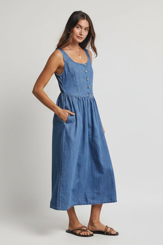 Woman wearing a medium blue denim tank dress against a gray 
background