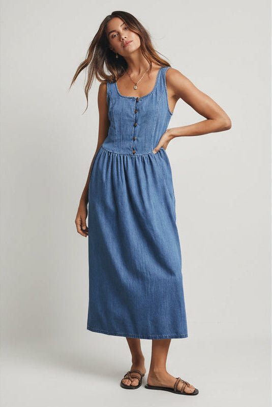 Woman wearing a medium blue denim tank dress against a gray 
background