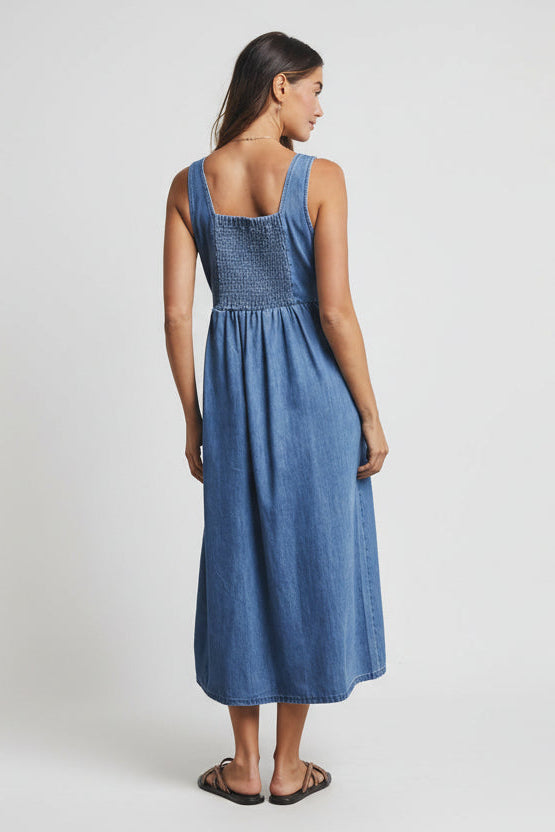 Back view of a woman wearing a medium blue denim tank dress with smocked back against a gray background