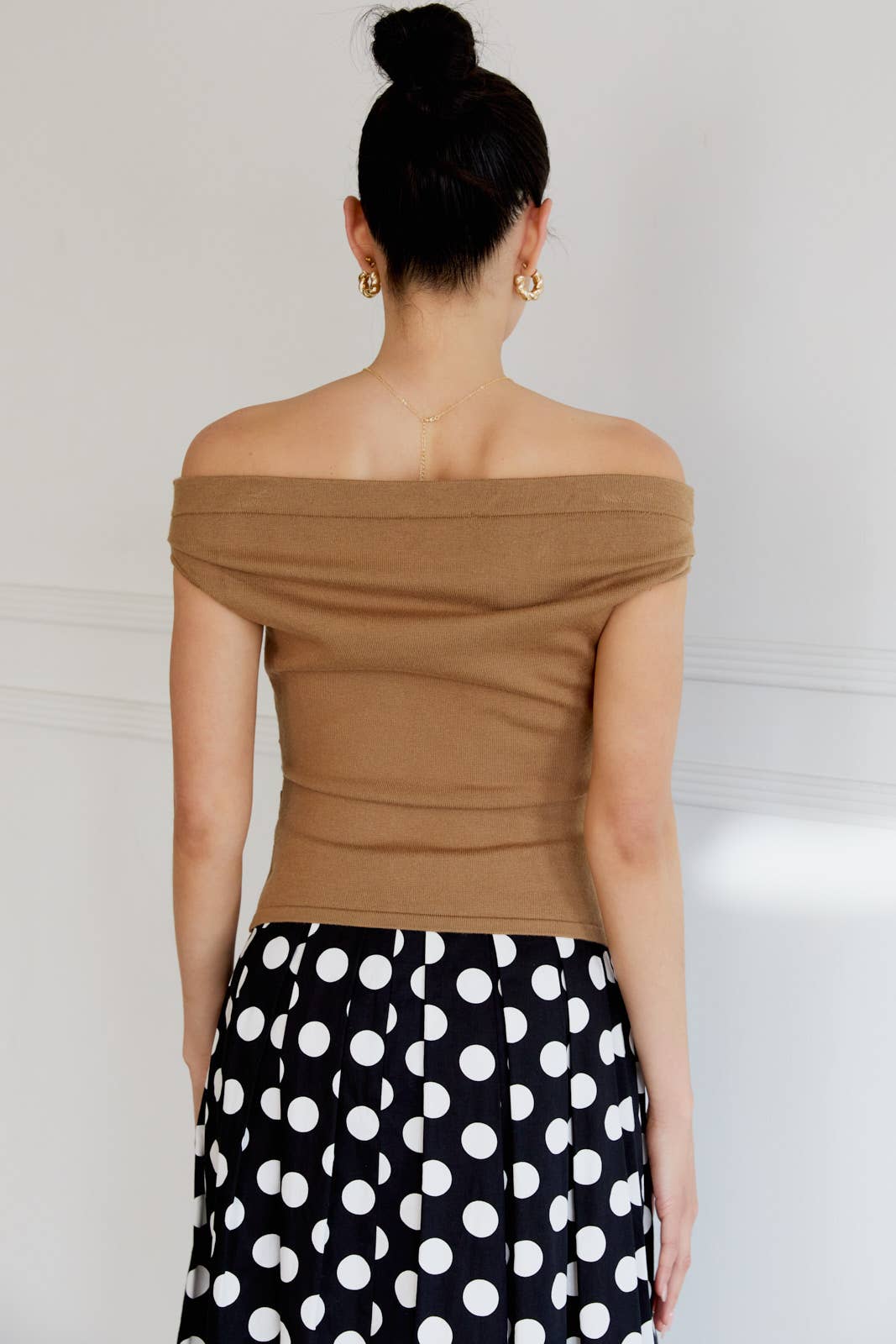 Back view of a woman wearing a tan off-shoulder top and black polka dot skirt against a white wall.