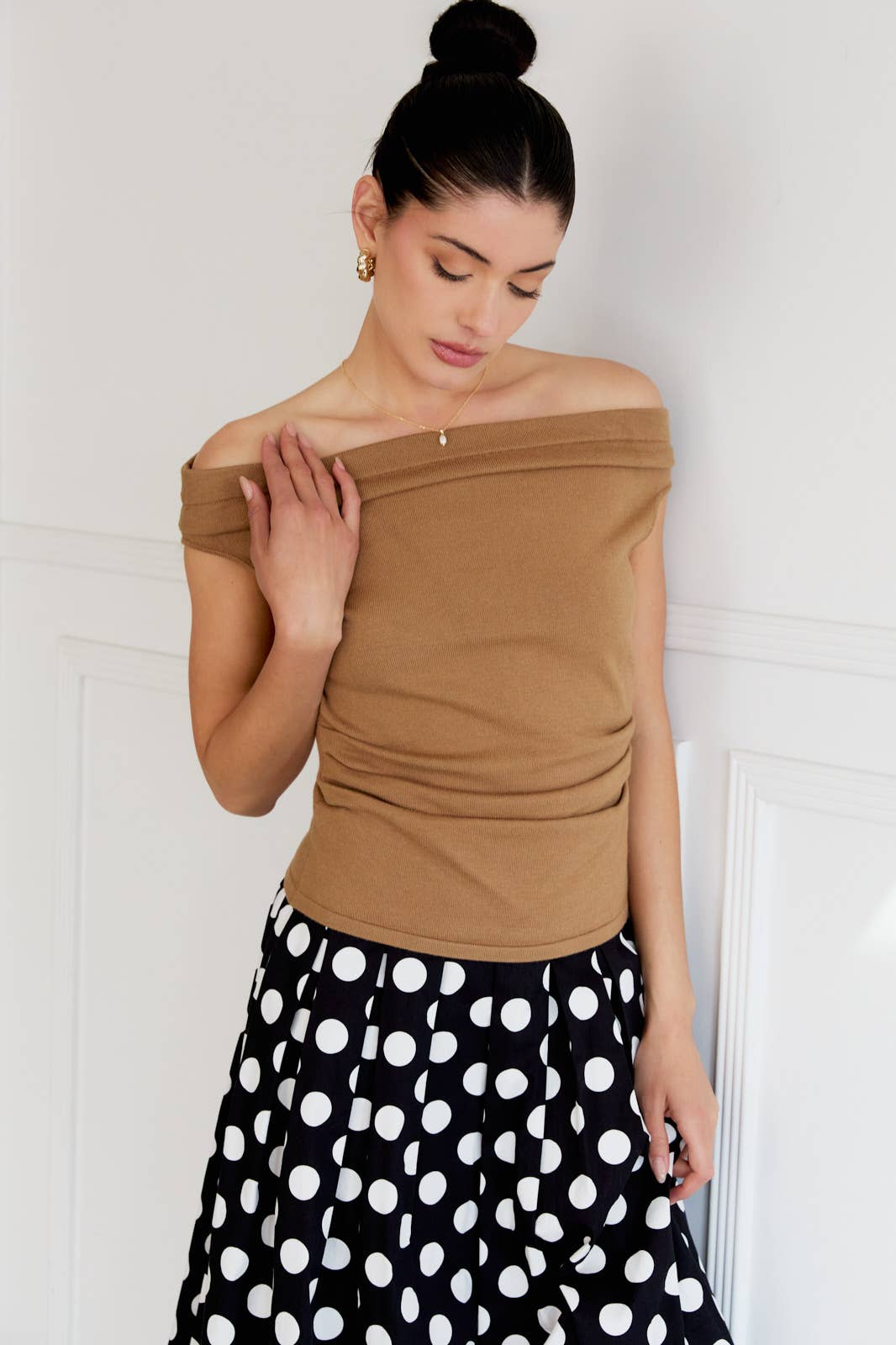 Woman wearing a tan off-shoulder top and black polka dot skirt against a white wall.