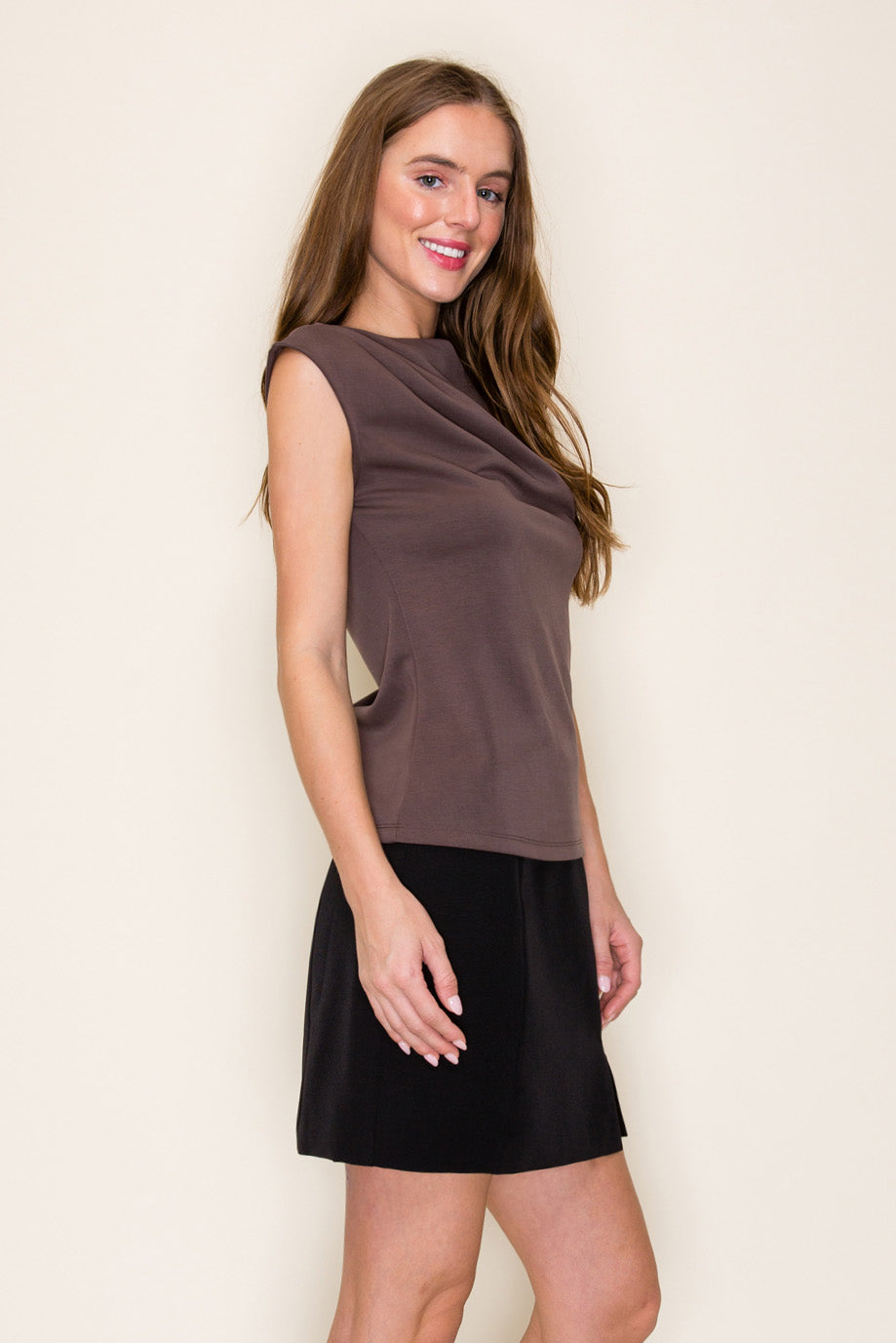 A woman modeling a brown scuba drape one shoulder top paired with a black skirt, standing against a plain off-white background.