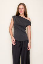 A woman modeling a dark grey scuba drape one shoulder modal top paired with black wide-leg pants, standing against a plain off-white studio background.
