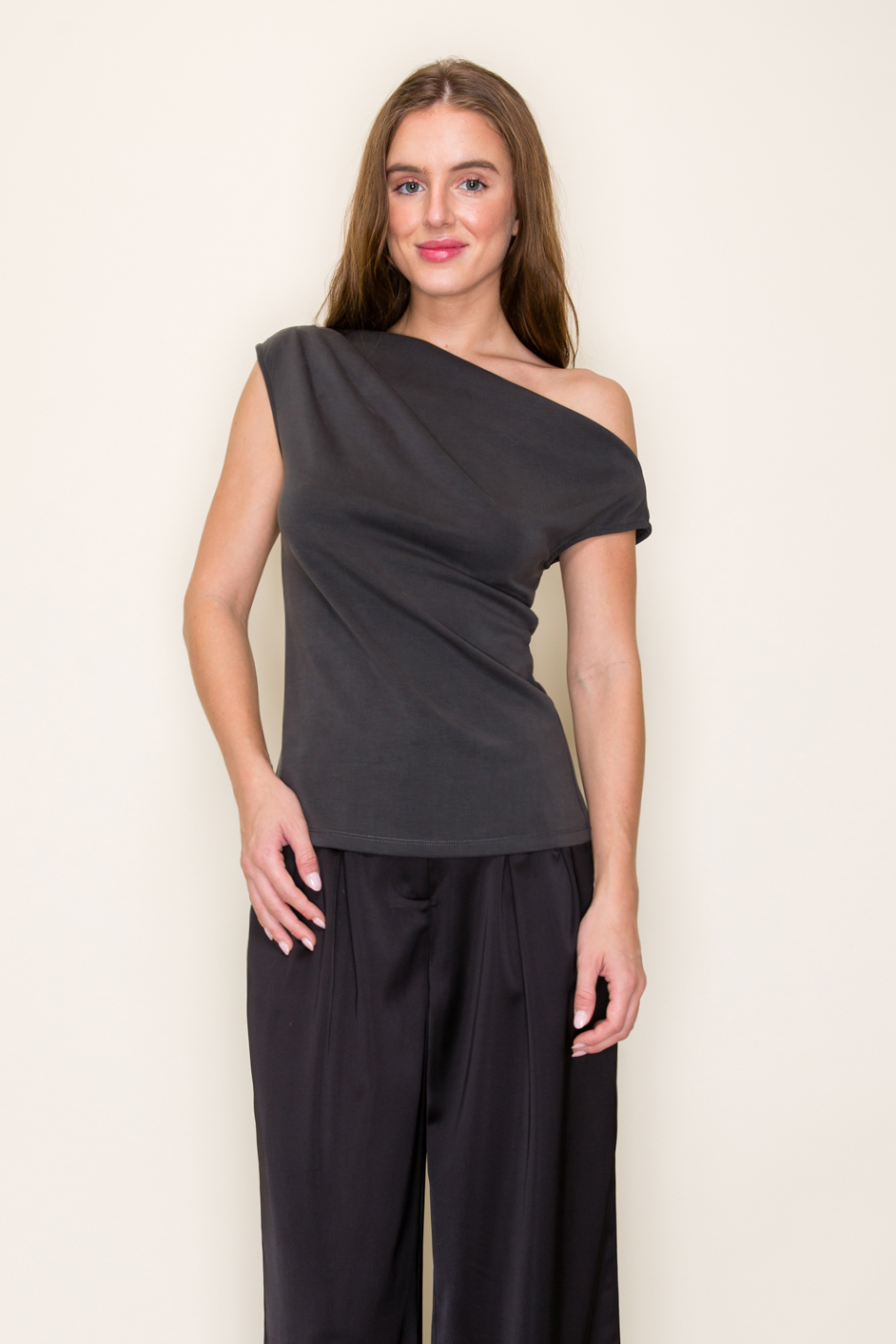 A woman modeling a dark grey scuba drape one shoulder modal top paired with black wide-leg pants, standing against a plain off-white studio background.