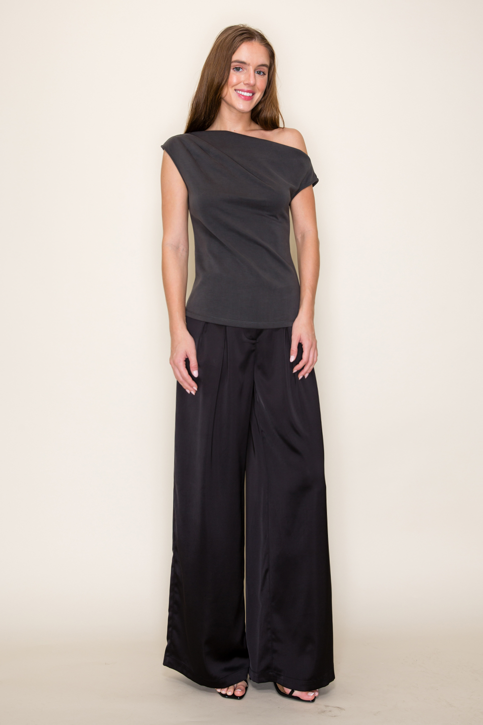 A woman modeling a dark grey scuba drape one shoulder modal top paired with black wide-leg pants, standing against a plain off-white studio background.
