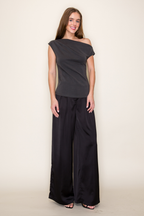 A woman modeling a dark grey scuba drape one shoulder modal top paired with black wide-leg pants, standing against a plain off-white studio background.