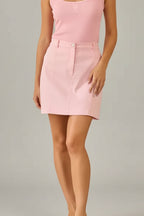 Woman wearing a pink tank top and skirt standing in a room with beige walls and curtains.