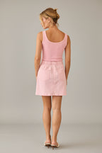 Back view of a woman wearing a pink tank top and skirt standing in a room with beige walls and curtains.