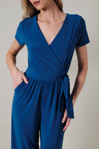 Woman wearing a royal blue faux wrap jumpsuit against a neutral background
