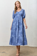 Woman wearing a blue jacquard tiered midi dress against a white wall.