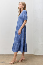 Woman wearing a blue jacquard tiered midi dress against a white wall.