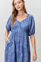 Woman wearing a blue jacquard tiered midi dress against a white wall.